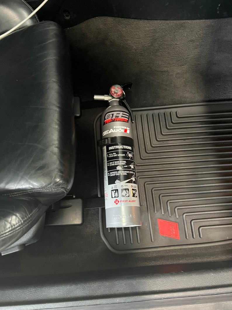 May include: A silver fire extinguisher with a black handle and a red gauge. The extinguisher is labeled "OFF-ROAD", "FIRE-TANK", "POWERED BY", "ODDAUGI", and "FIRST ALERT". The extinguisher is mounted in a car.