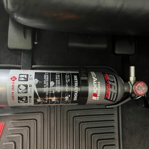 May include: A silver fire extinguisher with a black handle and a red gauge. The extinguisher is labeled "ME-TUNER" and "POWERED BY MODA AUDIO". The extinguisher is mounted in a car.
