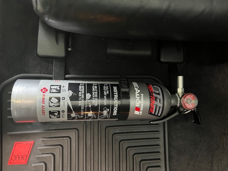 May include: A silver fire extinguisher with a black handle and a red gauge. The extinguisher is labeled "ME-TUNER" and "POWERED BY MODA AUDIO". The extinguisher is mounted in a car.