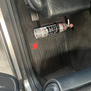 May include: A silver fire extinguisher is mounted to the floor of a car using a black strap. The extinguisher is labeled "First Alert" and has a red handle. The car has black floor mats with a red logo.