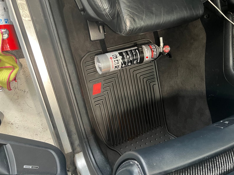 May include: A silver fire extinguisher is mounted to the floor of a car using a black strap. The extinguisher is labeled "First Alert" and has a red handle. The car has black floor mats with a red logo.