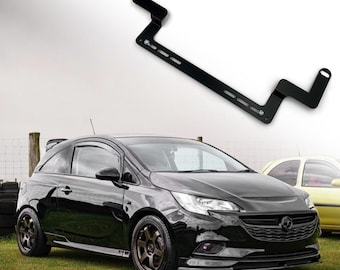 May include: A black, Z-shaped car number plate bracket is shown above a black coupe car on grass. The car has dark-coloured rims and a modern design. A yellow car is parked in the background.