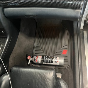 May include: A black car interior with a black floor mat and a fire extinguisher mounted on the side of the driver's seat. The fire extinguisher is silver with red and white lettering. The text on the fire extinguisher reads "First Alert 2.5A 10BC Fire Extinguisher".