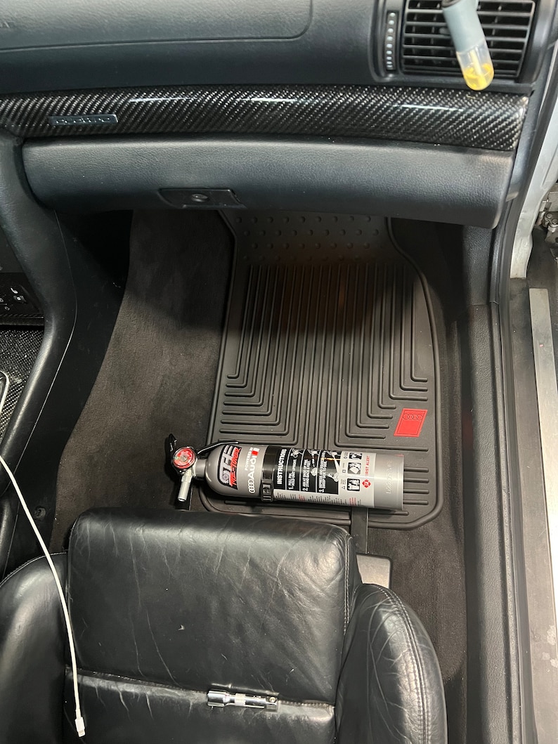 May include: A black car interior with a black floor mat and a fire extinguisher mounted on the side of the driver's seat. The fire extinguisher is silver with red and white lettering. The text on the fire extinguisher reads "First Alert 2.5A 10BC Fire Extinguisher".