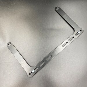 May include: Black metal bracket with multiple holes and two screws. The bracket is shaped like a rectangle with two arms extending out.