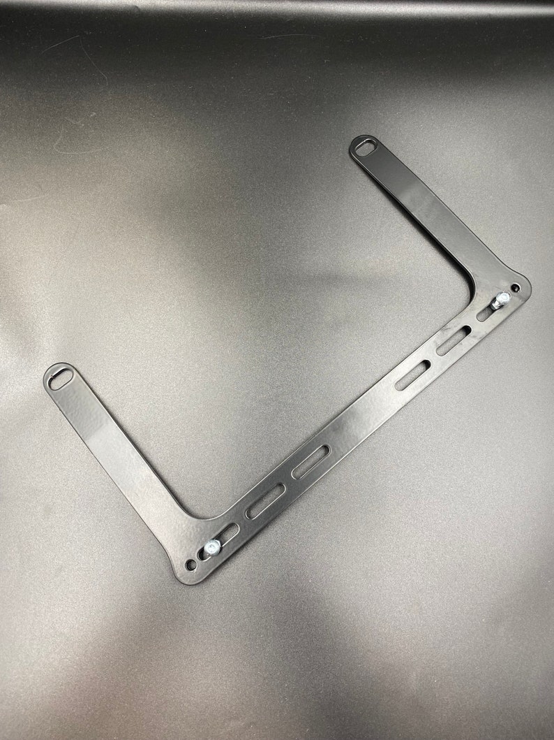 May include: Black metal bracket with multiple holes and two screws. The bracket is shaped like a rectangle with two arms extending out.
