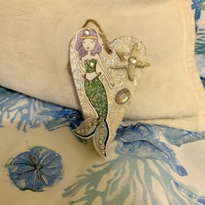 May include: A heart-shaped wooden ornament with a mermaid illustration. The mermaid has purple hair, a green tail, and is wearing a seashell bra. The ornament is decorated with glitter and a starfish.