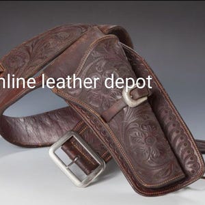 Puede incluir: Una funda de pistola de cuero marrón con intrincados diseños florales. La funda está unida a un cinturón de cuero marrón con una hebilla plateada. El texto "online leather depot" es visible en la imagen.