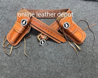 May include: A brown leather gun belt with two holsters and a silver buckle. The belt is decorated with stitching and the holsters have a silver emblem with a bird design. The text "online leather depot" is visible on the belt.