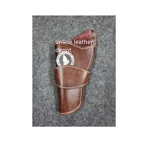 Könnte beinhalten: Ein braunes Lederholster mit weißen Nähten. Das Holster hat eine Klappe, die umgeklappt und mit einem Druckknopf befestigt wird. Das Holster hat ein Metallemblem mit dem Text "White Pigeon Depot" und einem weißen Taubenlogo.