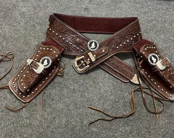 May include: A brown leather gun belt with two matching holsters. The belt has a silver buckle and decorative stitching. The holsters have silver accents and a small metal emblem.
