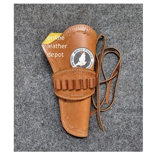 Könnte beinhalten: Ein braunes Lederholster mit einem Gürtelriemen und einem Riemen. Das Holster hat ein dekoratives Design und ein weißes Logo mit der Aufschrift "White Pigeon Depot".