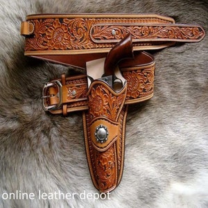 Op de afbeelding: Een bruine leren pistoolriem met ingewikkelde bloemmotieven en een bijpassende holster. De riem heeft een gesp met een zilveren afwerking. De holster is ontworpen om een ​​pistool te dragen. De riem en holster worden getoond op een bruine vacht achtergrond.