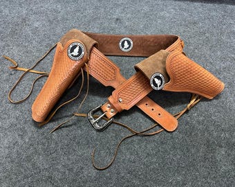 May include: A brown leather gun belt with two holsters and a silver buckle. The belt has a woven pattern and the holsters have a white bird logo with the text "White Pigeon Leather".