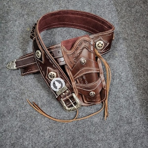 May include: A brown leather gun belt with a holster and a silver buckle. The belt features a star design and the holster has a Western-style design.
