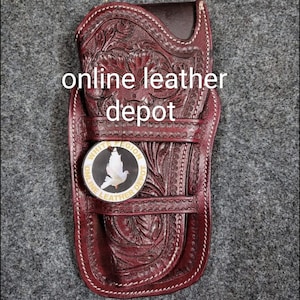 Puede incluir: Una funda de cuero marrón con intrincados detalles florales. La funda tiene una correa y una hebilla. Una etiqueta redonda dorada con el texto "White Pigeon Online Leather Depot" está unida a la funda.