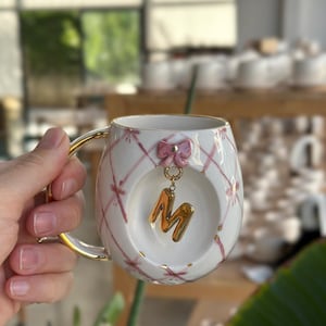Pode incluir: Caneca de cerâmica branca com alça e borda douradas, decorada com laços rosa e padrões cruzados. Um pingente dourado com a letra "M" pende da caneca. A caneca é segurada por uma pessoa.