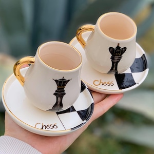 Puede incluir: Dos tazas de café de cerámica blanca con ribetes dorados y diseños de piezas de ajedrez negras. Cada taza tiene un platillo a juego con un patrón de tablero de ajedrez blanco y negro y la palabra "Chess" escrita en oro.