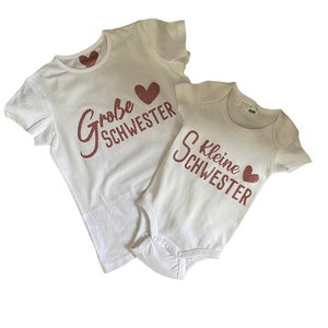 Puede incluir: Dos camisetas blancas con texto de purpurina rosa. La camiseta más grande dice "Große Schwester" con un corazón rosa. La camiseta más pequeña dice "Kleine Schwester" con un corazón rosa.