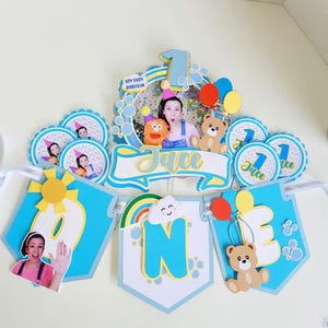 Puede incluir: Un conjunto de banderines azules y blancos para una celebración de primer cumpleaños. El banderín deletrea "ONE" con un sol, un arcoíris y un oso de peluche. El banderín también incluye una foto de una persona y el nombre "Jace".