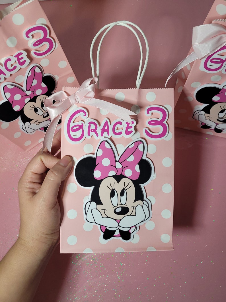 May include: Pink and white polka dot gift bag with a white handle. The bag has a Minnie Mouse character with a pink bow and white polka dots on her head. The bag also has a pink and white sticker that says "Grace 3".