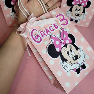 May include: Pink and white polka dot gift bag with a white handle and a pink bow. The bag features a Minnie Mouse character with a pink bow and white polka dots. The bag also has the name "Grace" written in pink letters with a pink number 3.