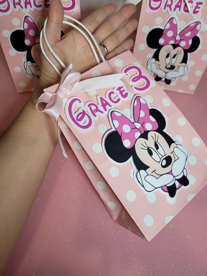 May include: Pink and white polka dot gift bag with a white handle and a pink bow. The bag features a Minnie Mouse character with a pink bow and white polka dots. The bag also has the name "Grace" written in pink letters with a pink number 3.