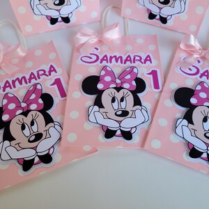 May include: Four pink and white polka dot gift bags with Minnie Mouse character cutouts. Each bag has a pink ribbon handle and a white label with the name "Samara" and the number "1" in pink.