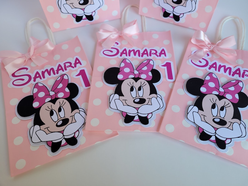 May include: Four pink and white polka dot gift bags with Minnie Mouse character cutouts. Each bag has a pink ribbon handle and a white label with the name "Samara" and the number "1" in pink.