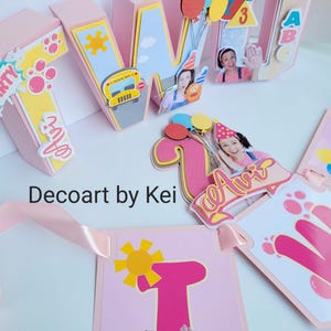 Puede incluir: Decoraciones de fiesta rosas y amarillas con las letras "T", "W", "A" y "1". Las decoraciones incluyen una pancarta, globos, un autobús escolar y un osito de peluche. También se ve el texto "Decoart by Kei".