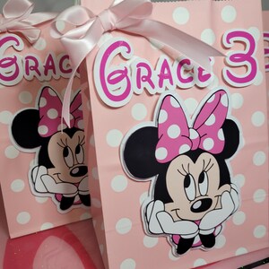 May include: Pink and white polka dot gift bags with a pink bow and a Minnie Mouse character. The bags are decorated with the name "Grace" and the number "3".