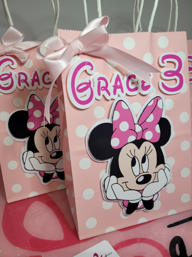 May include: Pink and white polka dot gift bags with a pink bow and a Minnie Mouse character. The bags are decorated with the name "Grace" and the number "3".