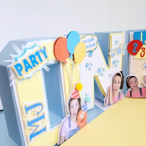 Puede incluir: Una exhibición de manualidades de papel con temática de fiesta con la palabra "PARTY" en estilo cómic, globos y las iniciales "MJ". La exhibición incluye recortes de personas y los números 2 y 3. La combinación de colores es azul, amarillo y naranja.