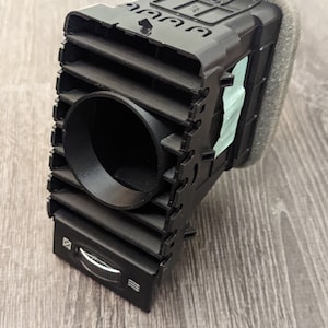 Puede incluir: Ventilación de aire de coche de plástico negro con una abertura redonda y una perilla plateada. La ventilación tiene una superficie texturizada y está diseñada para encajar en el tablero de instrumentos de un coche.
