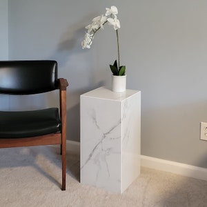 May include: A white marble pedestal with a white orchid plant in a white ceramic pot. The pedestal is sitting on a light grey carpet.