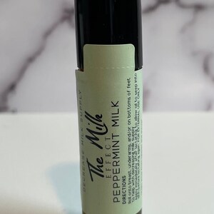 May include: A small, brown glass bottle with a black cap. The bottle has a label with the text "The Milk Effect Peppermint Milk." The label is light green with black text. The bottle is designed to help decrease milk supply.