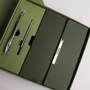 Puede incluir: Un diario de cuero verde con un cierre de metal dorado y la palabra "Simon" grabada en él. El diario está en una caja verde con dos bolígrafos.