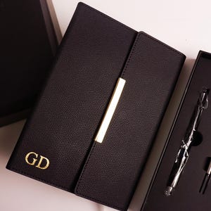 Op de afbeelding: Een zwart lederen notitieboek met een gouden penhouder en de initialen "GD" gegraveerd op de voorkant.