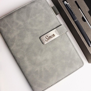 Peut inclure: Un carnet en cuir gris avec une fermeture en métal gravée du nom "Simon". Un stylo et un stylet assortis sont présentés dans un coffret cadeau. Le carnet est fermé et bordé de coutures.