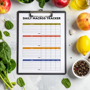 May include: A clipboard with a "DAILY MACROS TRACKER" form, surrounded by fresh produce. Red apples, spinach, a lemon, and a bowl of peppercorns are visible. The form has sections for breakfast, lunch, and dinner, with columns for protein, carbs, fats, and calories.