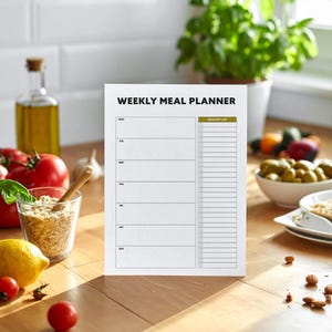 May include: A white weekly meal planner with the text "WEEKLY MEAL PLANNER" at the top. The planner has sections for each day of the week and a grocery list. The planner is on a wooden surface.
