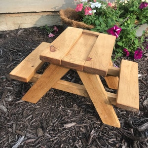 May include: A small wooden picnic table with two benches, stained in a light brown color. The table is sitting on a bed of brown wood chips.