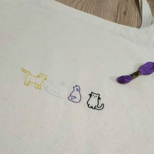 Peut inclure: Un sac fourre-tout blanc avec quatre chats brodés de différentes couleurs : jaune, blanc, violet et noir. Les chats sont alignés et regardent vers la droite du sac. Une bobine de fil à broder violet se trouve dans le coin supérieur droit de l’image.