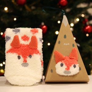 May include: A pair of fuzzy white socks with a fox face design, packaged in a brown cardboard box shaped like a Christmas tree. The box has a small fox face on the front.