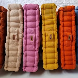 Puede incluir: Seis cojines de suelo cuadrados en diferentes colores, incluyendo naranja, marrón, rosa, amarillo, beige y azul. Cada cojín tiene un diseño acolchado y una etiqueta de cuero marrón con la palabra "MEJA" impresa en ella.