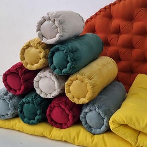 May include: A stack of colourful cylindrical pillows in various shades of red, yellow, green, and grey. The pillows are arranged in a pyramid shape on a yellow cushion.