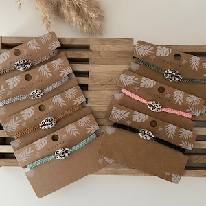 Könnte beinhalten: Fünf Armbänder mit Leopardenmuster-Muschel-Anhängern auf braunen Karten mit weißen tropischen Blattdesigns und dem Wort "MUSTHAVE" auf jeder Karte.