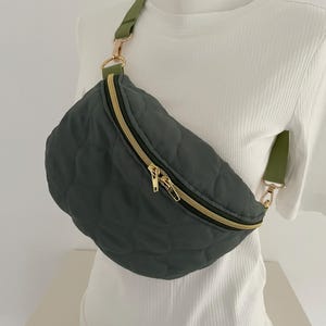 Könnte beinhalten: Gesteppte, dunkelgrüne Bauchtasche mit goldenem Reißverschluss und olivgrünem Riemen. Die Tasche hat eine runde Form und wird quer über dem Körper getragen. Der Riemen ist verstellbar und wird mit goldfarbenen Metallverschlüssen befestigt.