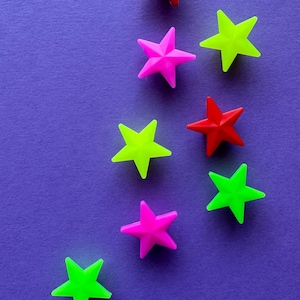 Könnte beinhalten: Eine Ansammlung von acht kleinen, fünfzackigen sternförmigen Objekten, die in einem verstreuten Muster angeordnet sind. Die Sterne sind in leuchtenden Farben wie Rot, Neonpink und Neongrün gehalten und bilden einen Kontrast zur violetten Fläche.