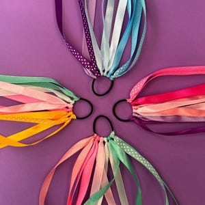 Könnte beinhalten: Vier schwarze Haargummis, die jeweils mit einem Bündel mehrfarbiger Satinbänder verziert sind. Die Bänder zeigen verschiedene Schattierungen von Lila, Blau, Pink, Orange, Gelb und Grün, wobei einige mit weißen Punkten gemustert sind.